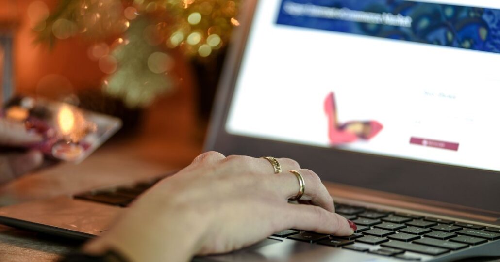 A hand typing on a laptop showing an online shoe store, with warm lights behind