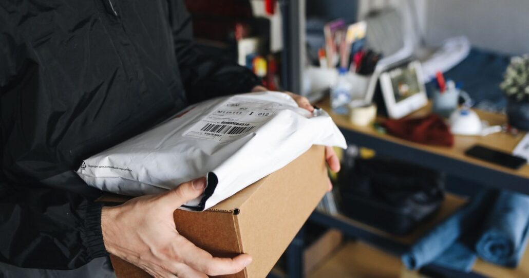 A courier holding a padded mailer and a shipping box in a workshop