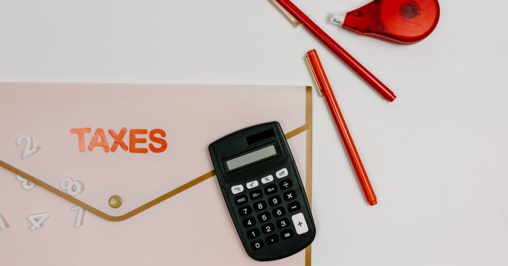 An envelope marked taxes beside a calculator and pens on a pink background